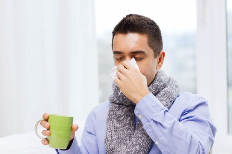 Healthcare, flu, people, rhinitis and medicine concept - ill man blowing his nose with paper napkin and drinking tea at home