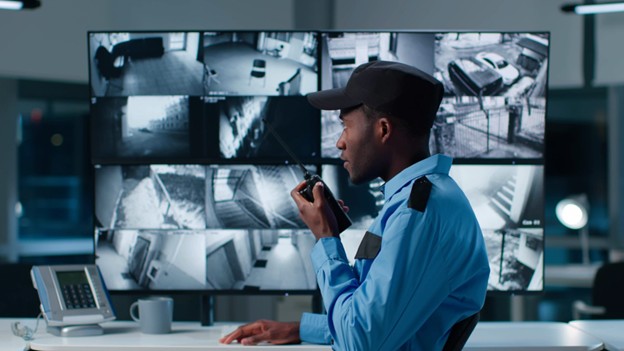 A security guard in uniform holds a walkie-talkie while monitoring multiple surveillance camera feeds displayed on large screens in a control room.