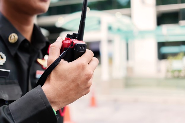A person in a uniform holds a walkie-talkie, appearing to communicate outdoors as part of client-centric security. Their face is partially out of frame, with blurred buildings and traffic cones seen in the background.