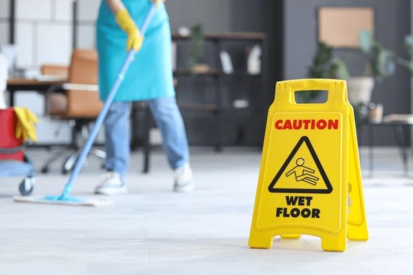 A yellow "Caution: Wet Floor" sign is placed on the floor, while a person in gloves and an apron mops in the background of an office or commercial space.
