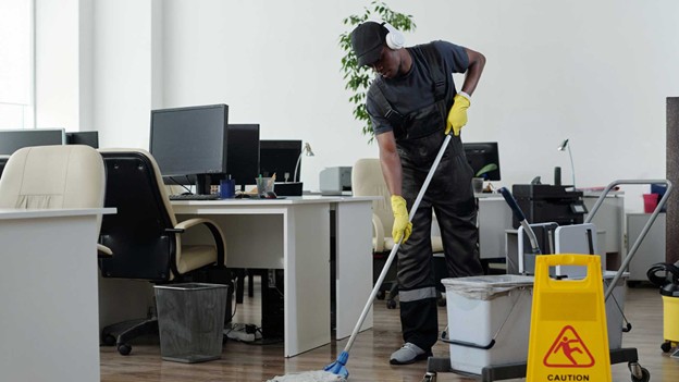 A person wearing headphones, gloves, and work overalls is mopping the floor in an office with desks and computers. A yellow "Caution: Wet Floor" sign is placed nearby. Cleaning equipment is visible.