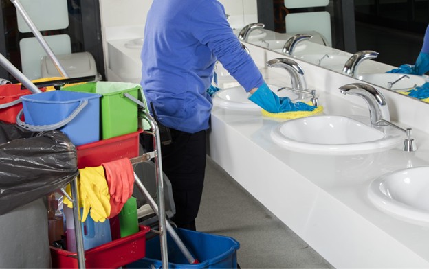 A person wearing blue gloves and a blue shirt cleans a sink in a public restroom. Nearby is a cleaning cart with buckets, cleaning supplies, and rubber gloves hanging over the side.