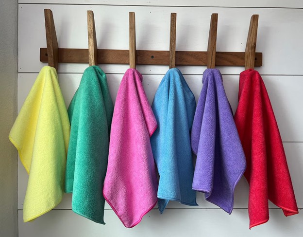 Five colorful towels (yellow, green, pink, blue, and red) hang side by side from wooden pegs on a white wall.