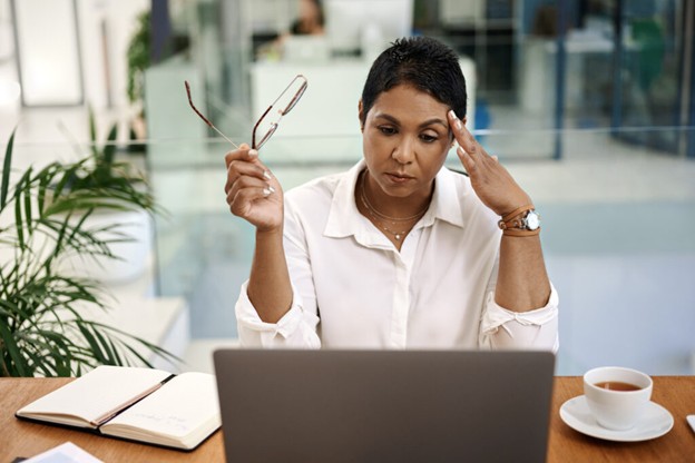 A woman sits at a desk with a laptop, holding her glasses in one hand and touching her temple with the other, appearing stressed or deep in thought. An open notebook and a cup of coffee are on the table.