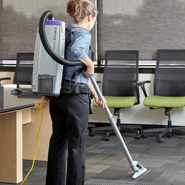 A person wearing a backpack vacuum cleaner is cleaning a carpeted office space with several empty chairs and desks in the background.