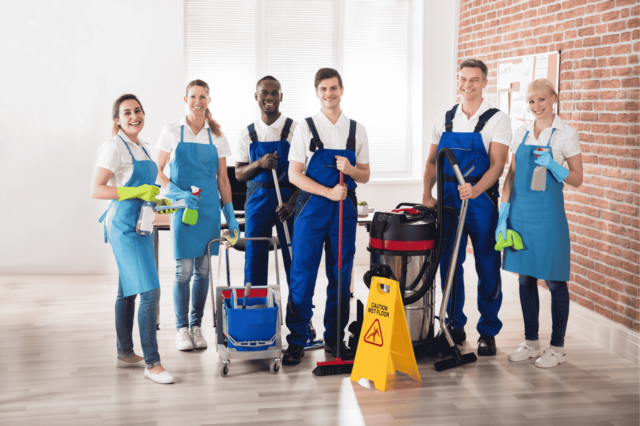 A group of six smiling janitors in blue uniforms stand together with cleaning supplies, including a mop, bucket, vacuum, and caution sign, in a bright, modern room with wooden floors and brick walls.