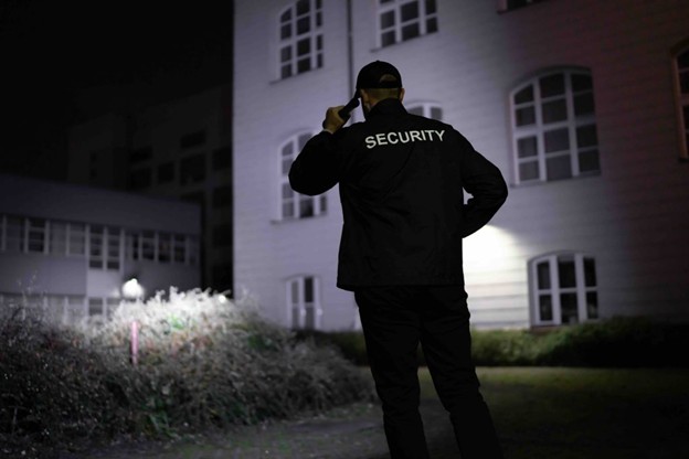 A security guard in a black uniform and cap stands outside a building at night, talking on a walkie-talkie. The word "SECURITY" is printed on the back of the jacket. The scene is dimly lit.